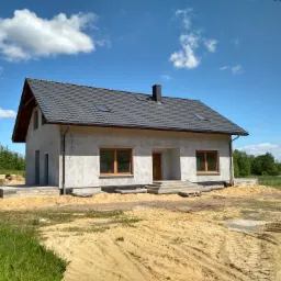 Surowy dom jednorodzinny z ciemną dachówką i drewnianymi oknami, w trakcie budowy, z widocznymi śladami opon na piaszczystym terenie.