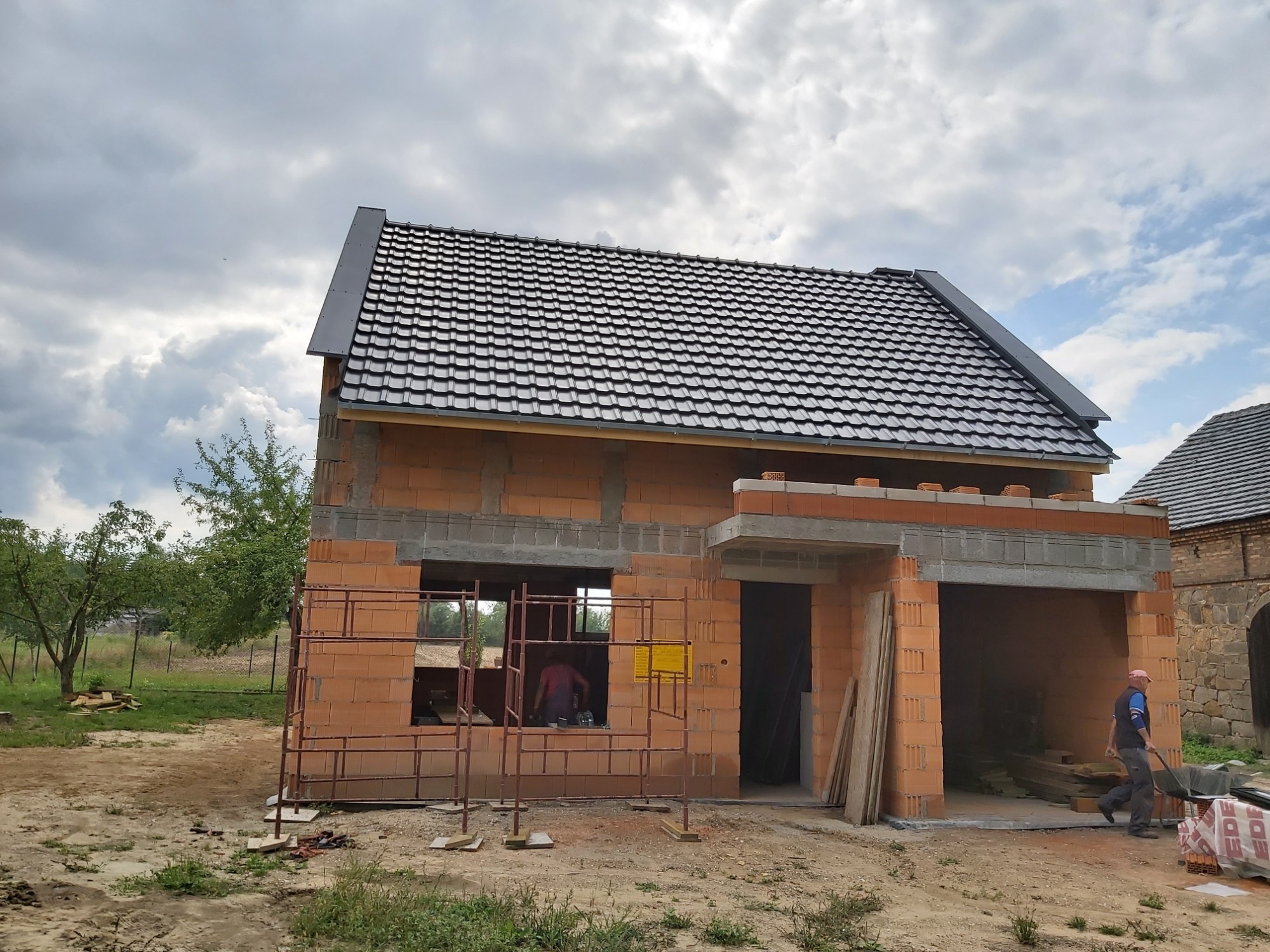 Surowy stan domu murowanego z ciemną dachówką, widoczne rusztowania i pracownik z taczką, etap budowy w słoneczny dzień.