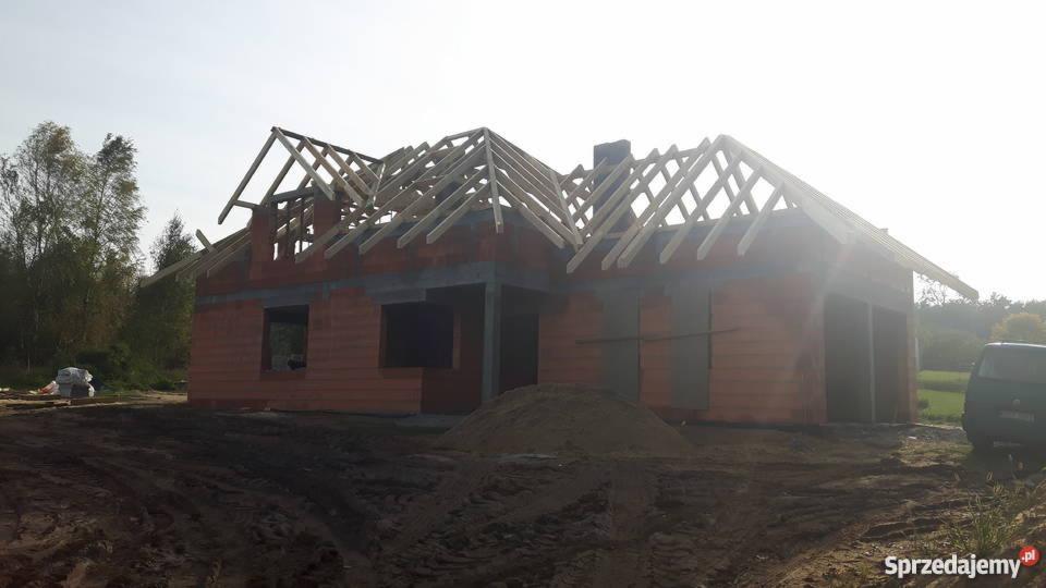 Surowy dom murowany w trakcie budowy, widoczne ściany z czerwonej cegły, drewniana konstrukcja dachu, sterta piasku i teren budowy z widocznymi śladami opon pojazdu.