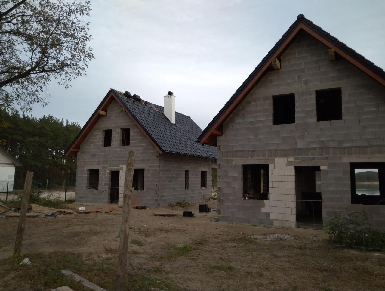 Dwa domy jednorodzinne w stanie surowym zamkniętym z ciemną dachówką, widoczne ślady murowania z bloczków betonowych, jeden z kominem, w tle las i jezioro.