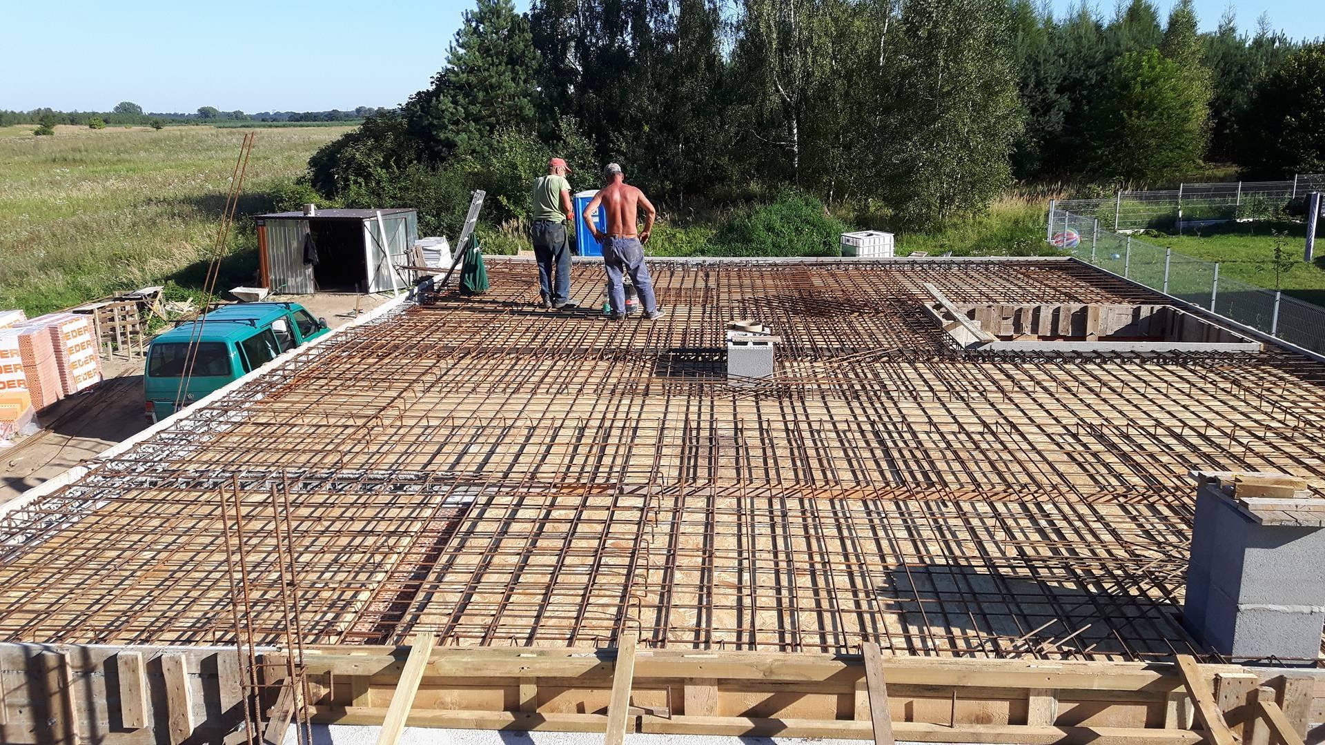 Zbrojenie płyty fundamentowej domu jednorodzinnego z widocznymi pracownikami, szalunkiem, starym busem VW i stosem cegieł w tle.
