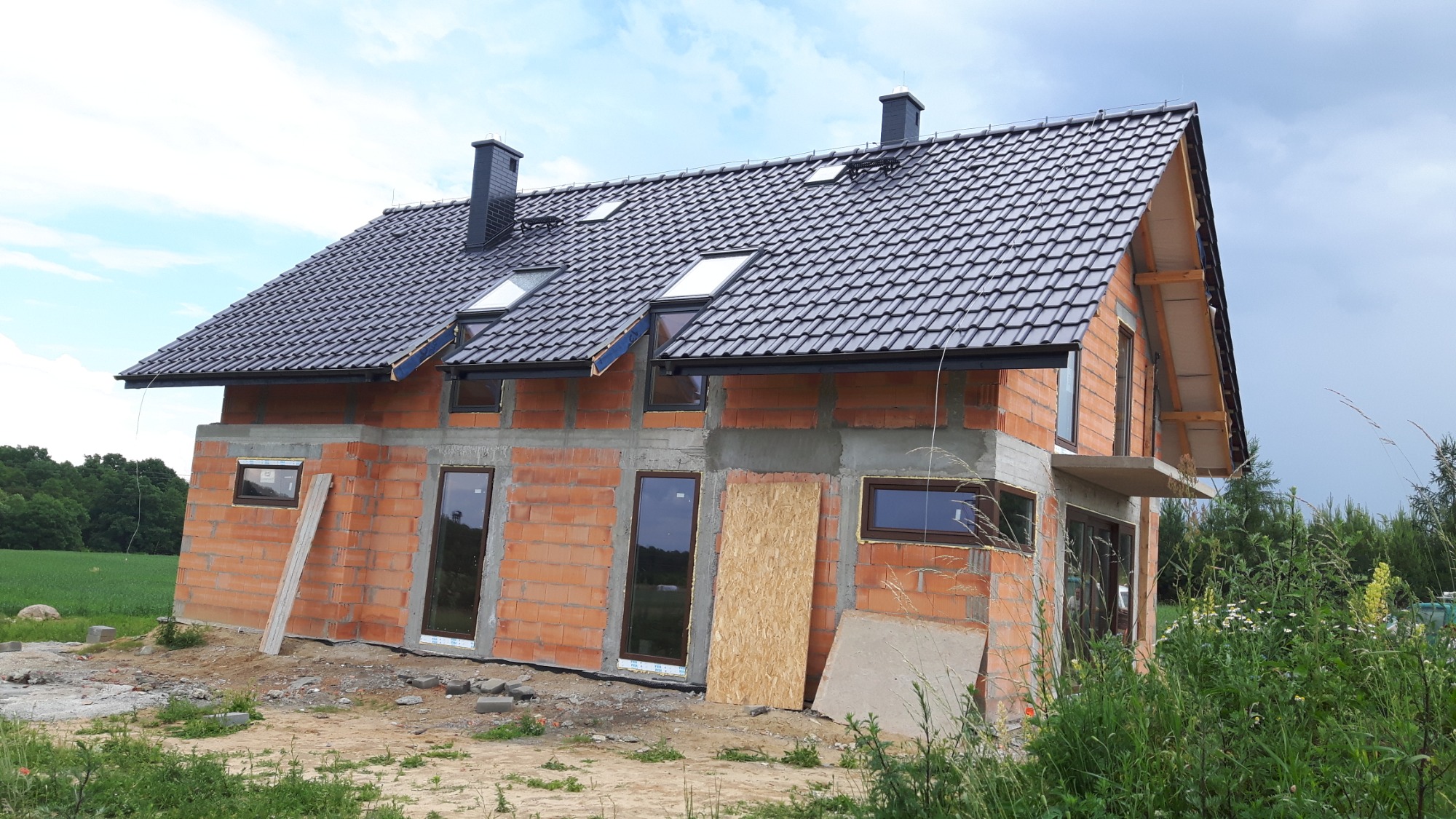 Surowy stan domu z czerwonej cegły z czarnym dachem z dachówki, widoczne okna i kominy, na tle zieleni i pochmurnego nieba.