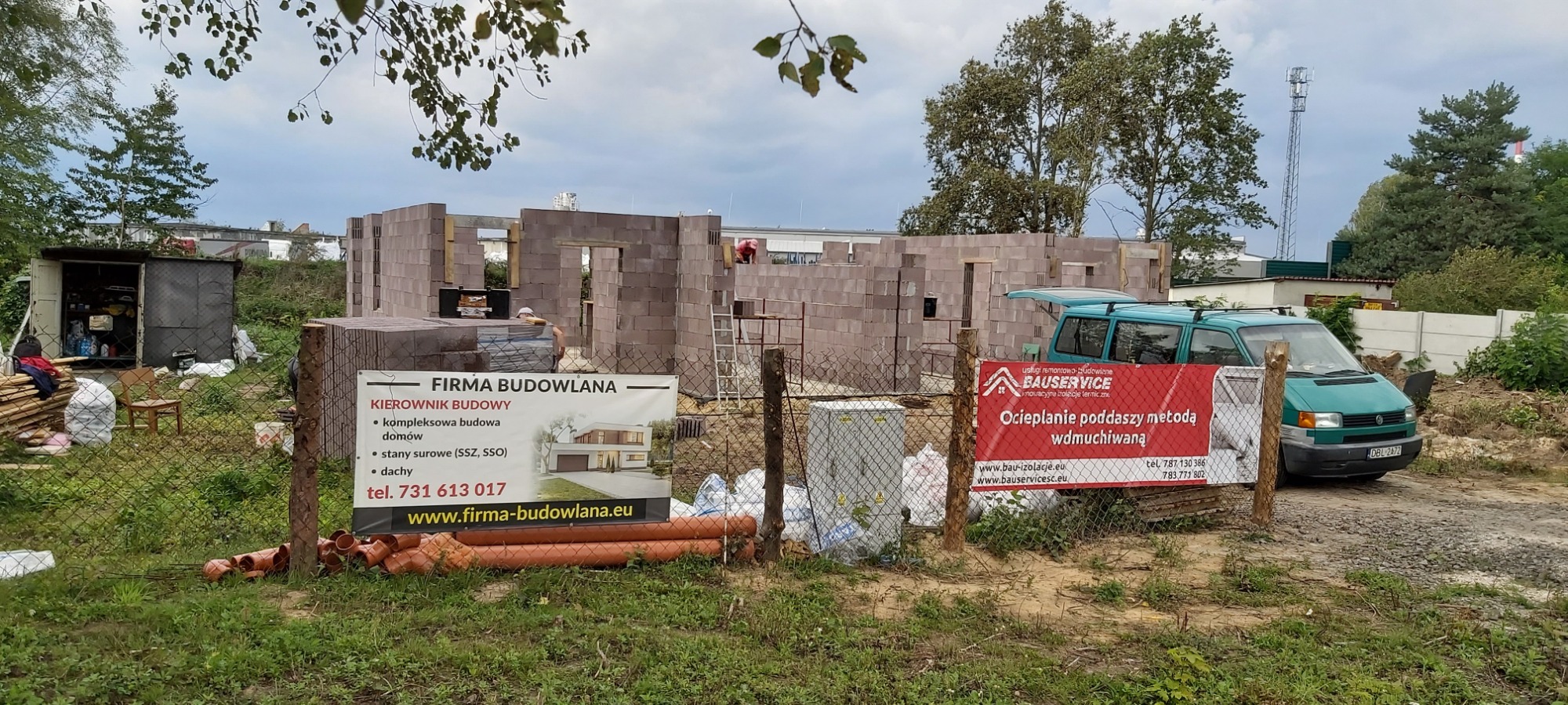 Budowa domu w stanie surowym, widoczne ściany z bloczków, tablice reklamowe firm budowlanych przy ogrodzeniu, zielony bus w tle.