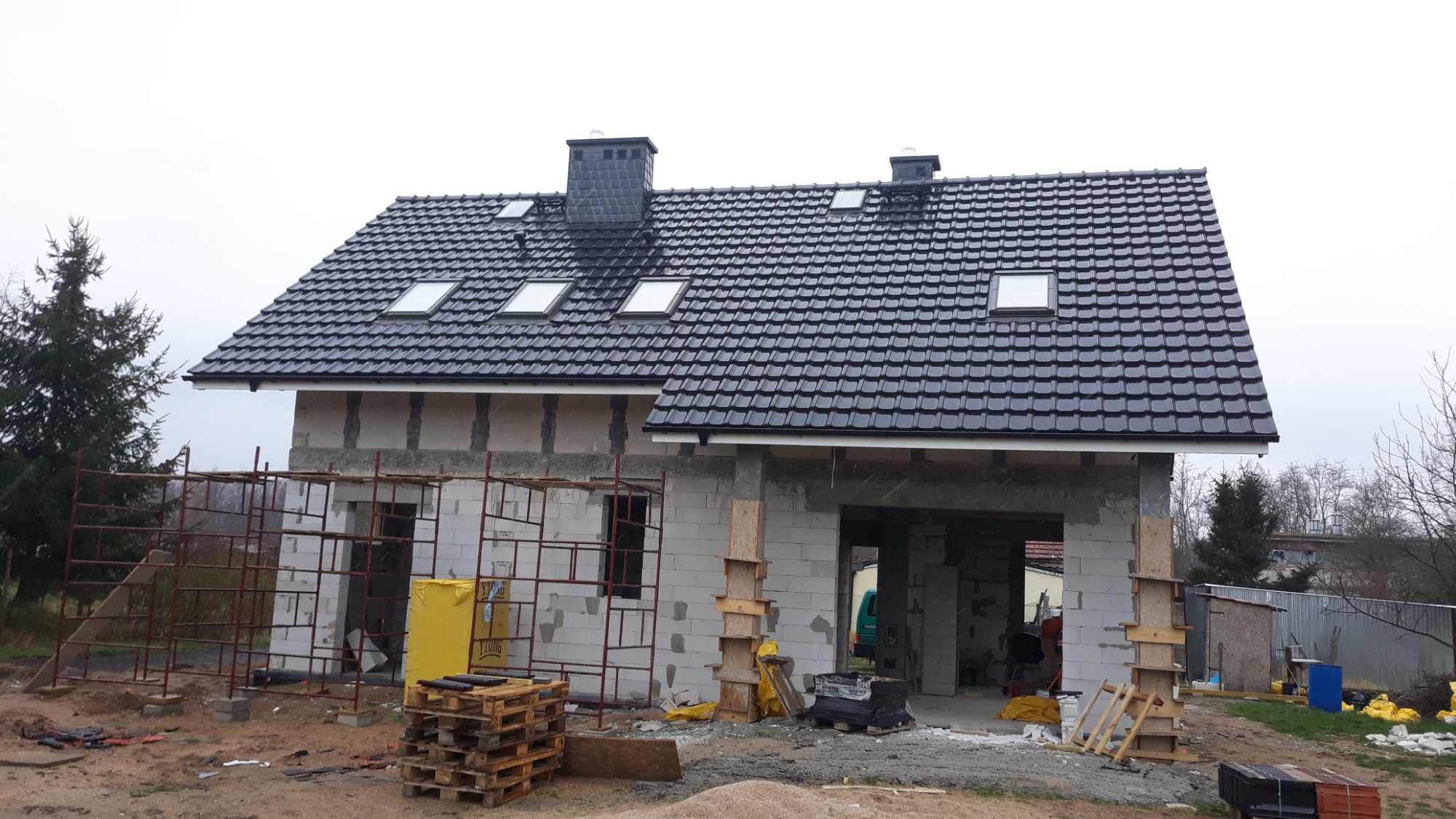 Dom w trakcie budowy, ściany z betonu komórkowego, czarny dach z dachówką ceramiczną i oknami połaciowymi, widoczne rusztowania i materiały budowlane.