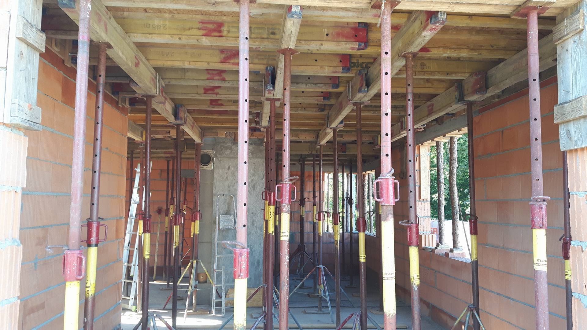 Wnętrze domu w trakcie budowy, widoczne murowane ściany z czerwonej cegły, drewniane szalunki stropowe podparte metalowymi stemplami budowlanymi, drabina w tle.