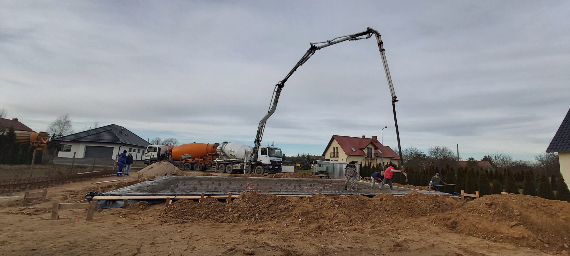 Wylewanie betonu na zbrojenie fundamentów domu jednorodzinnego przy użyciu pompy do betonu, widoczne dwie betoniarki i pracownicy w trakcie pracy.