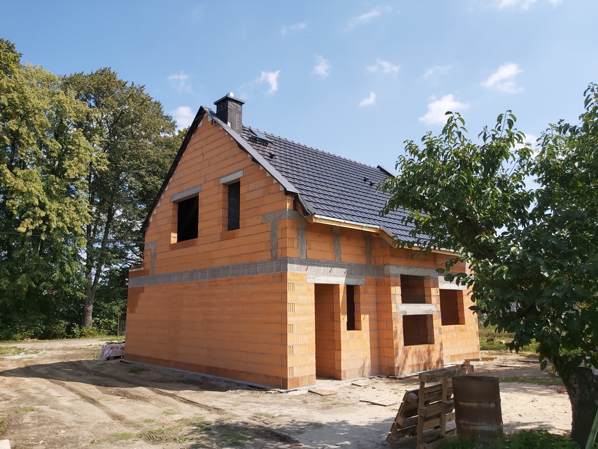 Niedokończony, dwukondygnacyjny dom z czerwonej cegły z ciemnoszarą dachówką, widoczne otwory okienne i drzwiowe, komin z czarną obudową, drzewa w tle, paleta i beczka na pierwszym planie.