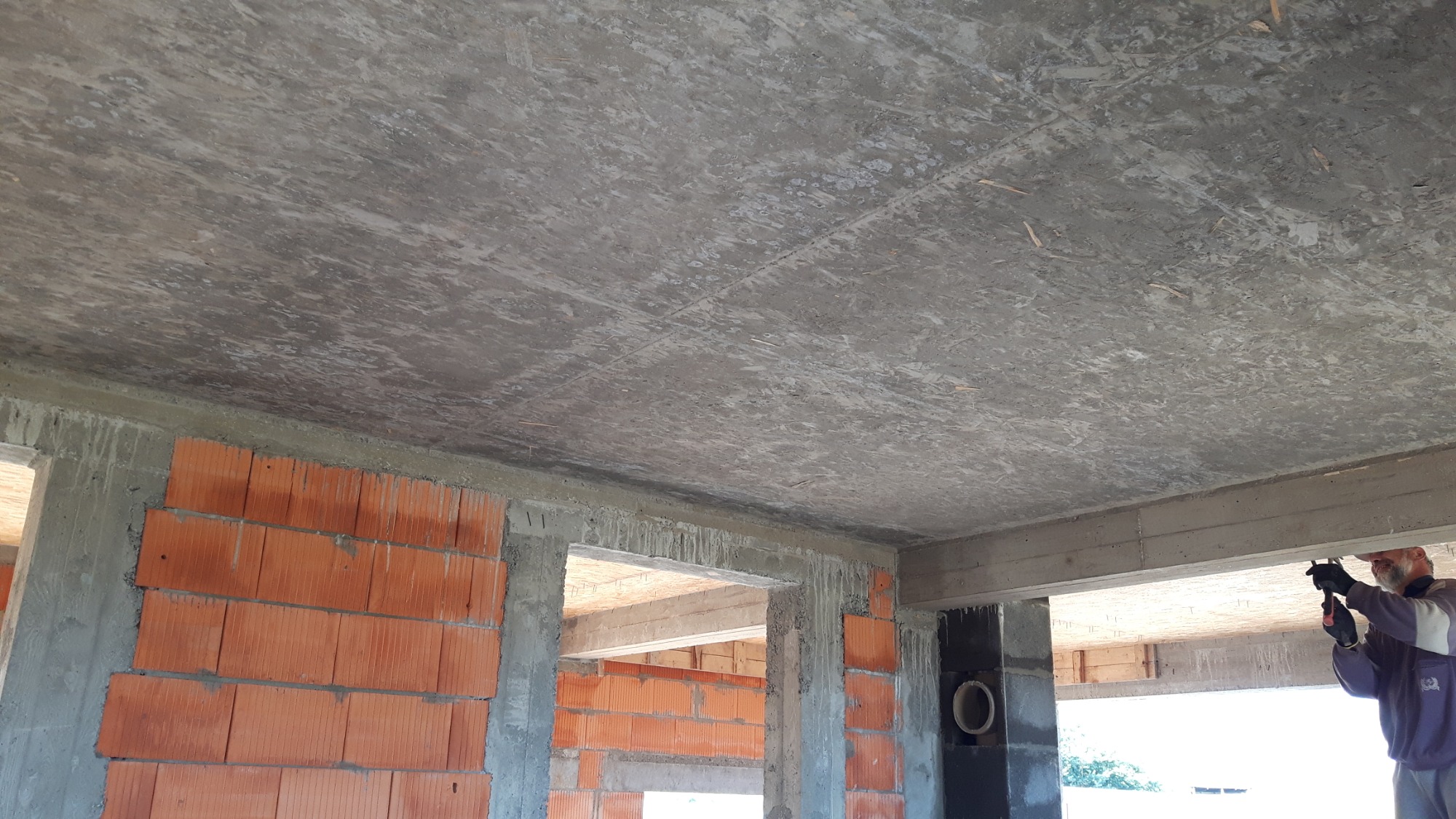 Surowy strop z widoczną strukturą desek szalunkowych w nowo wznoszonym budynku z czerwonej cegły, fragment pracującego murarza w prawym dolnym rogu.