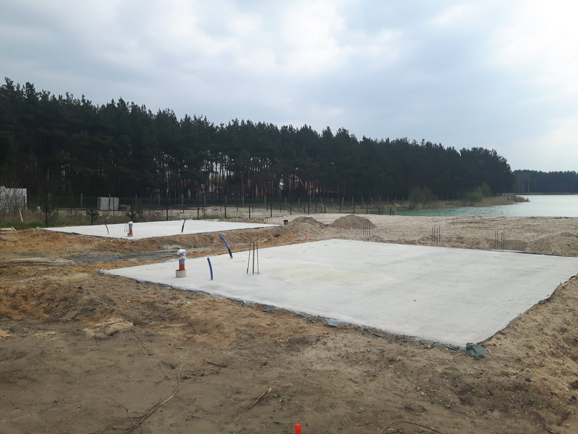Dwie betonowe płyty fundamentowe na piaszczystym terenie, widoczne wystające rury i pręty zbrojeniowe, w tle las i brzeg jeziora pod pochmurnym niebem.