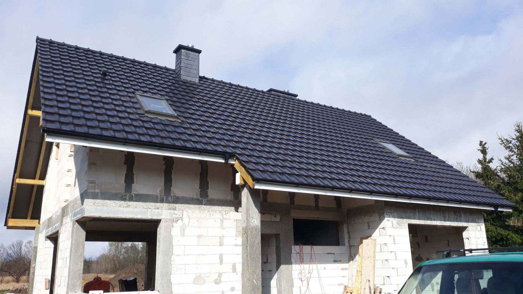 Niedokończony dom murowany z czarną dachówką, widoczne okna dachowe i komin, ściany z betonu komórkowego z widocznymi spoinami, fragment samochodu w prawym dolnym rogu.