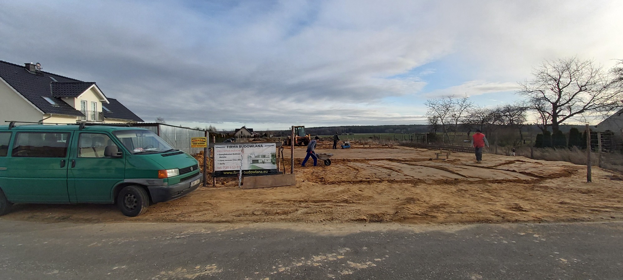 Przygotowanie terenu pod budowę domu murowanego: widok na wykopane fundamenty, pracujących robotników z taczkami, mini koparkę i tablicę informacyjną firmy budowlanej na tle pochmurnego nieba...