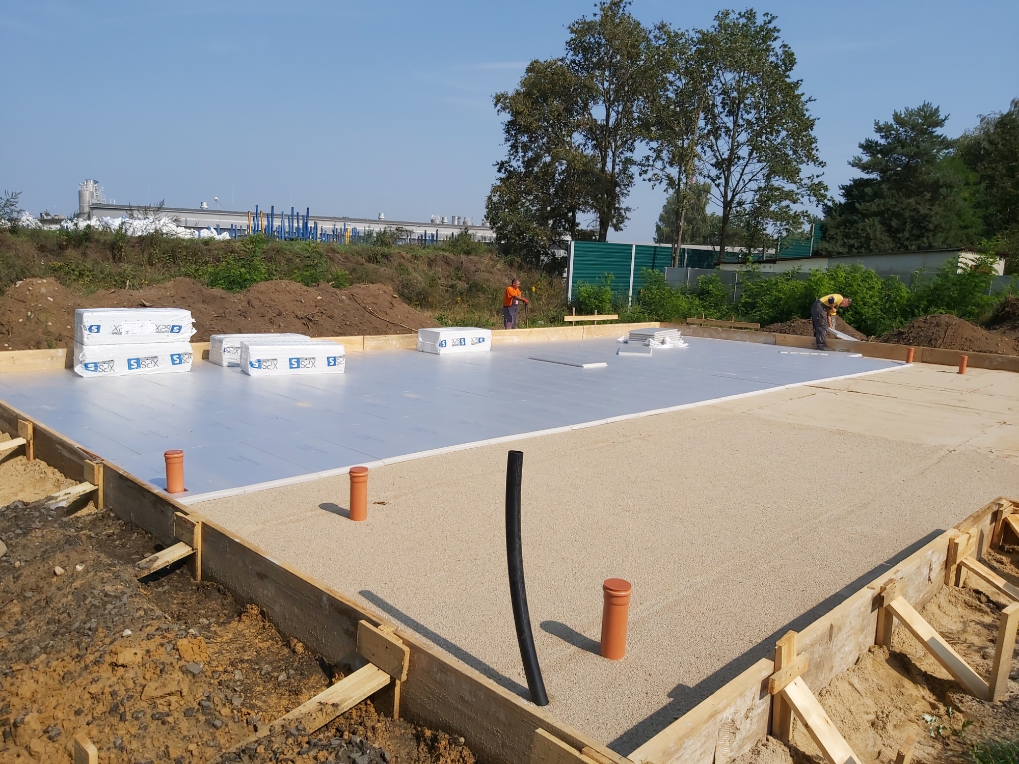 Przygotowanie płyty fundamentowej: deskowanie, izolacja termiczna XPS i podsypka piaskowa, dwóch pracowników w tle.