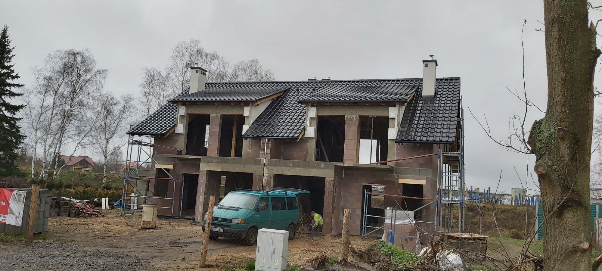 Dwukondygnacyjny dom murowany w trakcie budowy, z widoczną więźbą dachową pokrytą czarną dachówką, niezadaszonymi otworami okiennymi i drzwiami garażowymi, zielony bus VW Transporter zaparkowany...