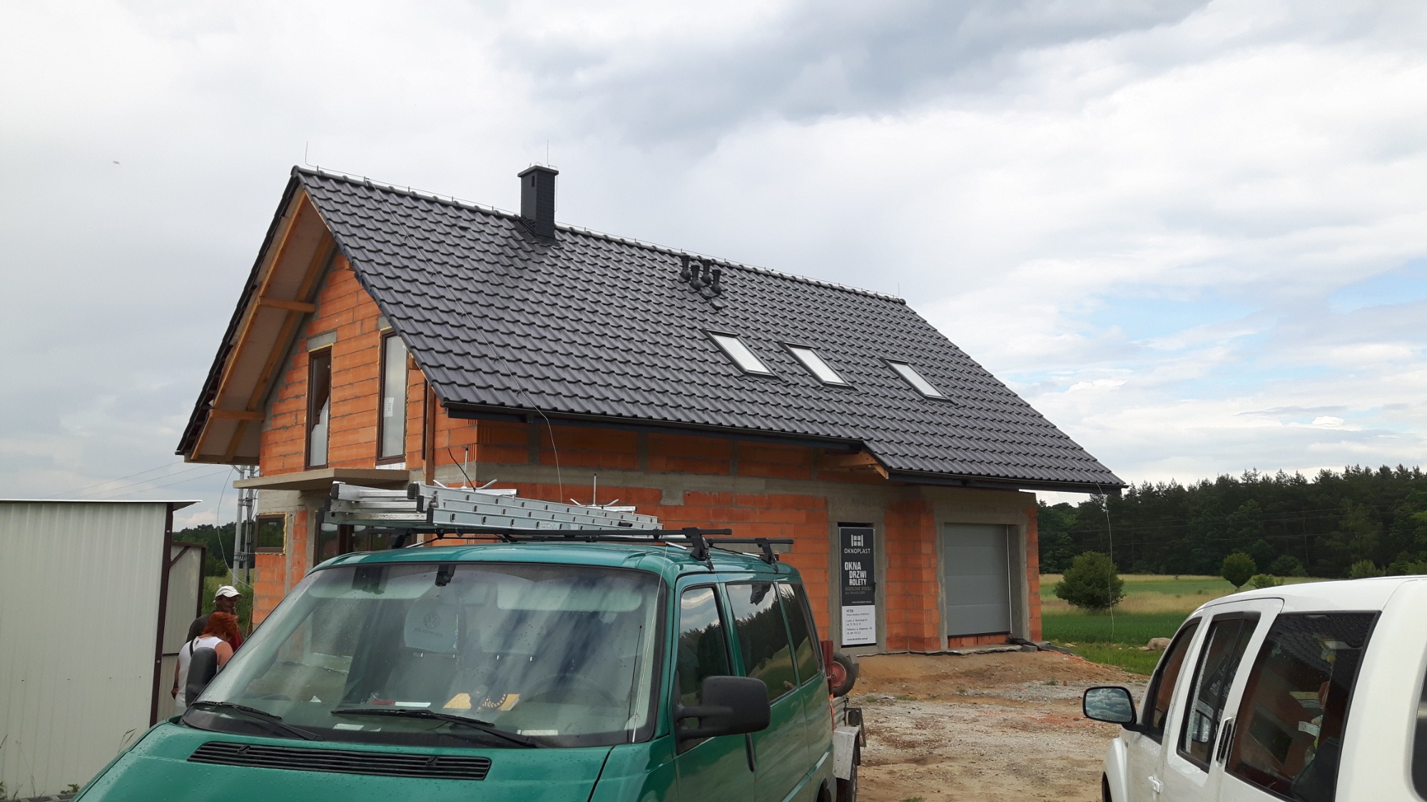 Dom murowany w trakcie budowy, z widoczną więźbą dachową, pokryty czarną dachówką z oknami dachowymi, na dachu zamontowany komin, na pierwszym planie zielony bus z drabiną na dachu, częściowo...