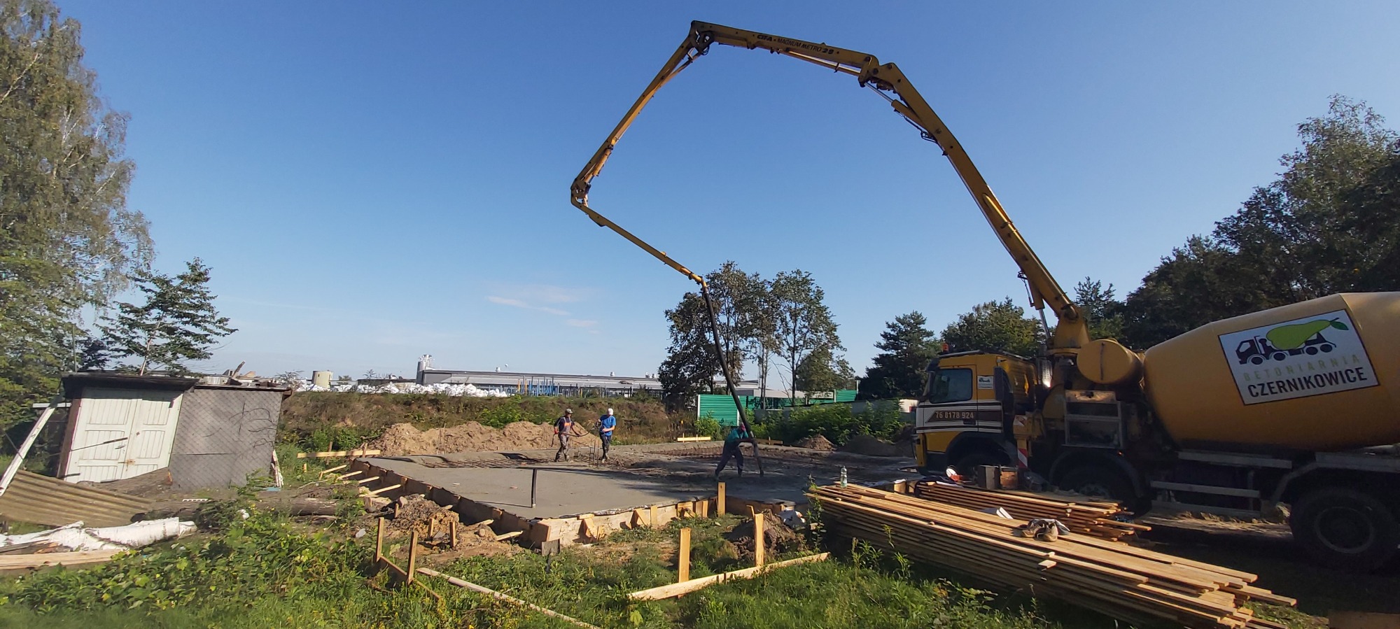 Wylewka fundamentów domu jednorodzinnego z użyciem pompy do betonu, widoczni pracownicy w trakcie rozprowadzania betonu, w tle mikser betonu z logo firmy Betoniarnia Czernikowice.