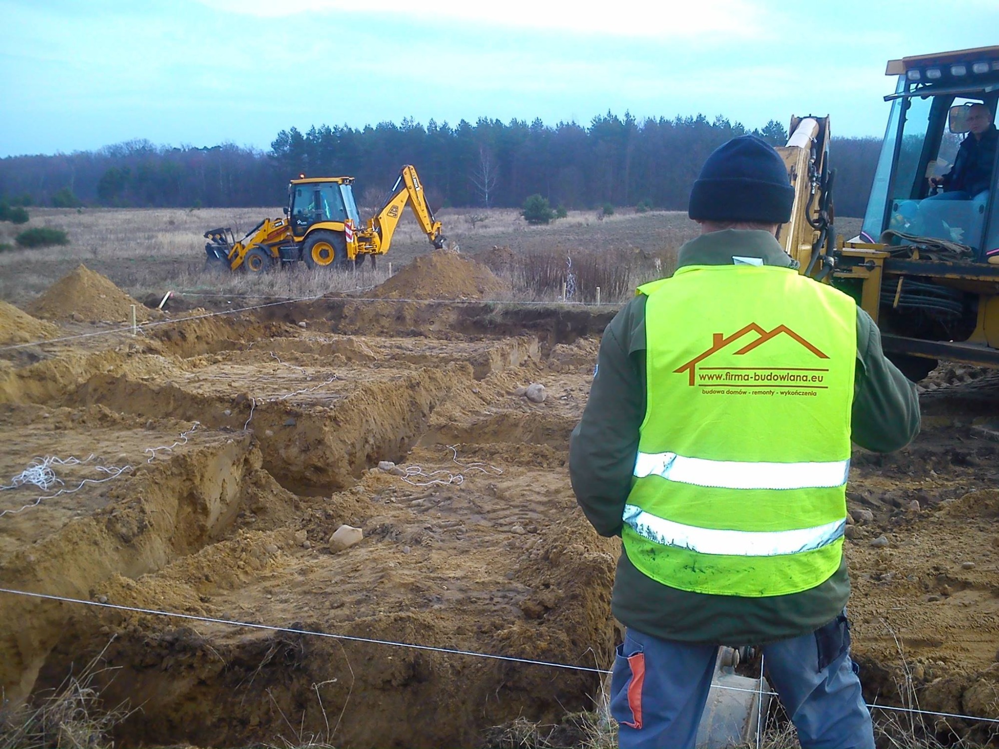 Przygotowanie terenu pod budowę: wykop fundamentów, pracująca koparka, pracownik w kamizelce odblaskowej obserwuje postęp prac.