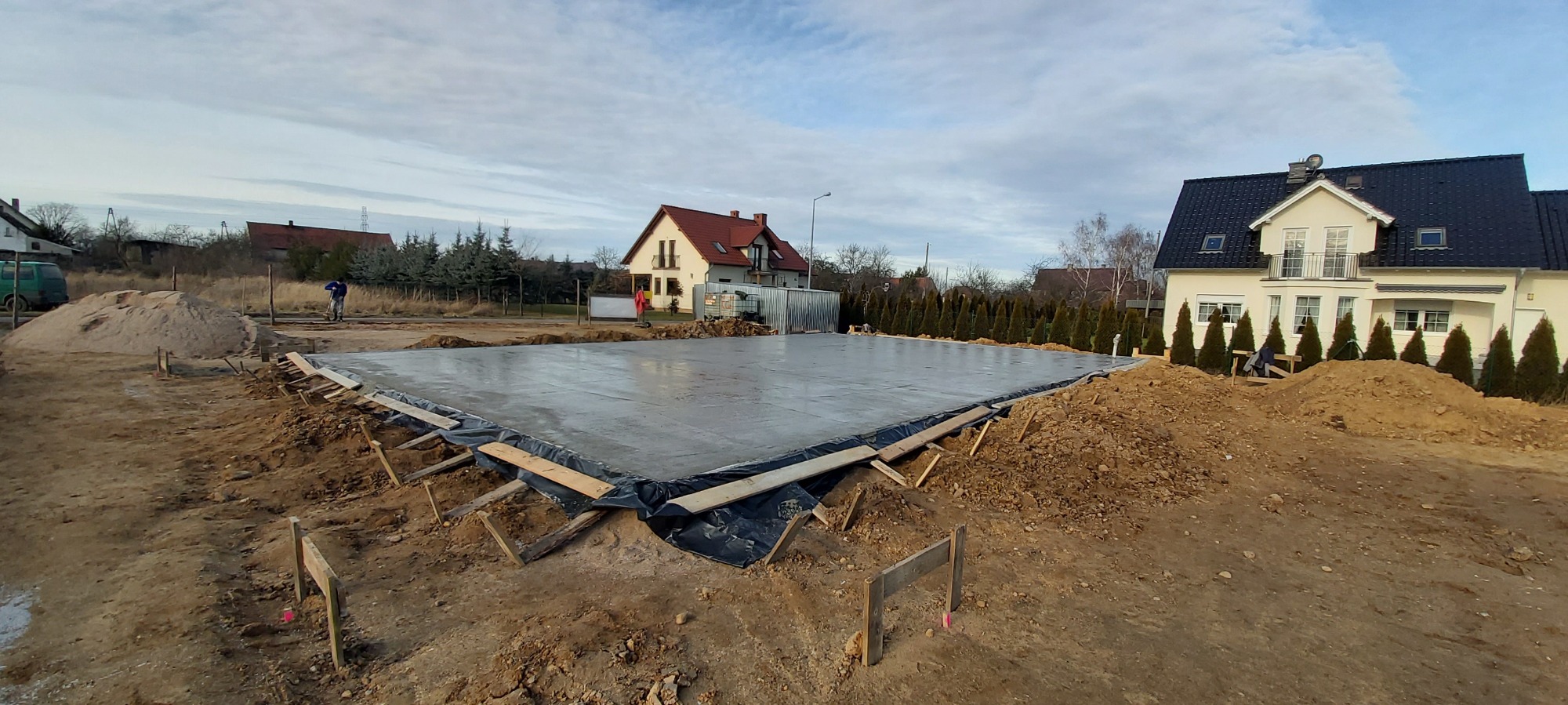 Wylana płyta fundamentowa pod dom jednorodzinny, zabezpieczona folią, widoczne drewniane szalunki i hałdy ziemi, w tle zabudowa jednorodzinna i błękitne niebo z chmurami.