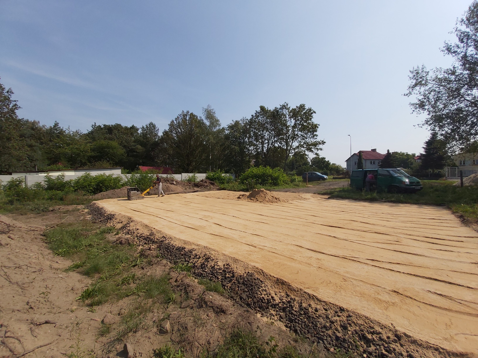 Przygotowany teren pod budowę, wyrównany piasek z widocznymi śladami po maszynie, w tle drzewa i budynki.