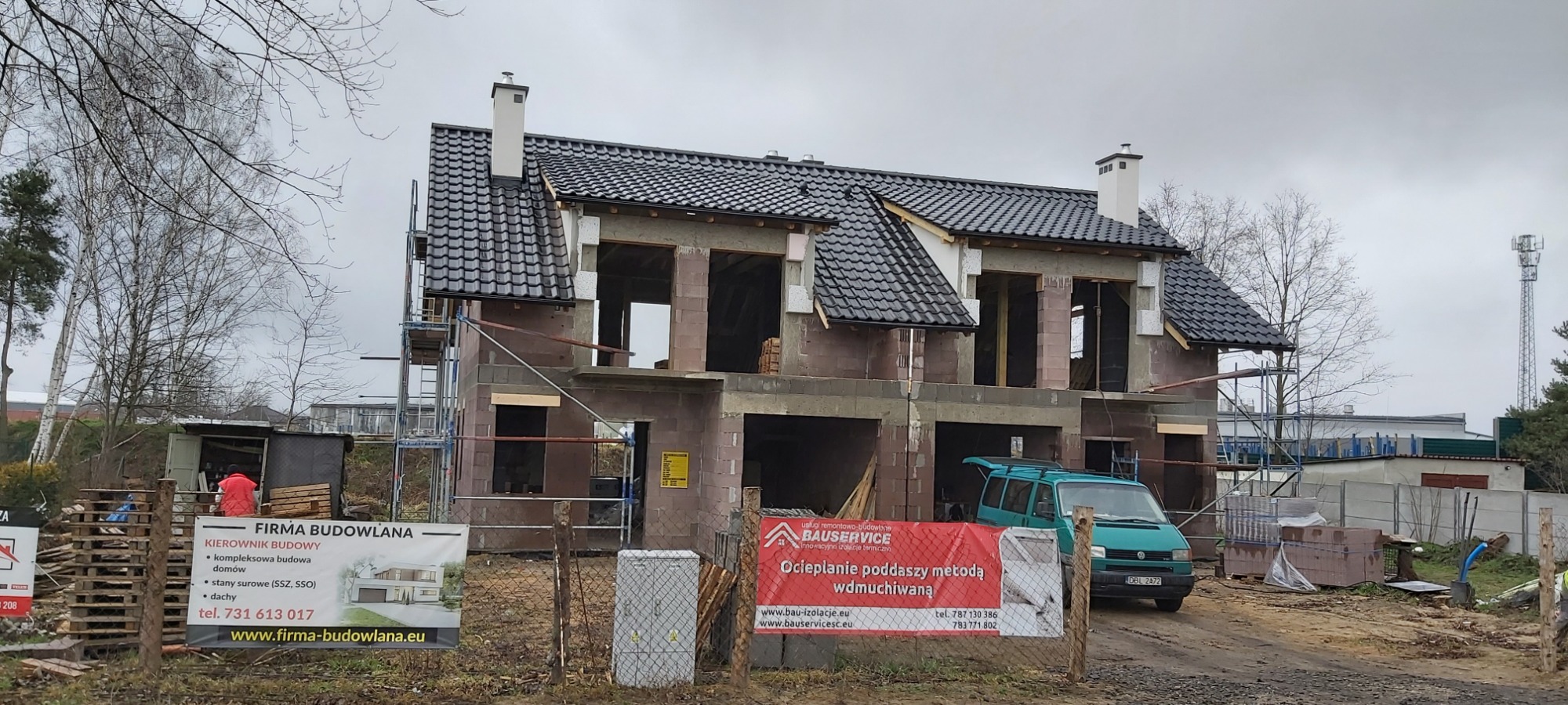Dwukondygnacyjny dom w trakcie budowy, widoczne surowe mury z pustaków, czarny dach z dachówki, rusztowania, reklamy firm budowlanych w tle, szara pogoda.