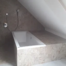 Biała wanna w zabudowie z płytek imitujących kamień, zamontowana w łazience na poddaszu ze skośnym sufitem i oknem dachowym.