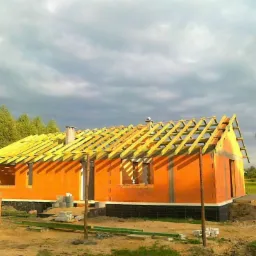 Dom murowany w stanie surowym otwartym z widoczną więźbą dachową, wykonany z pomarańczowych bloczków i izolacją fundamentów, na tle pochmurnego nieba.