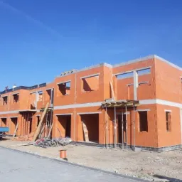 Wznoszenie ścian z czerwonej cegły porotherm w dwukondygnacyjnym budynku w stanie surowym otwartym, widoczni pracownicy na dachu, niebieska ciężarówka w tle.