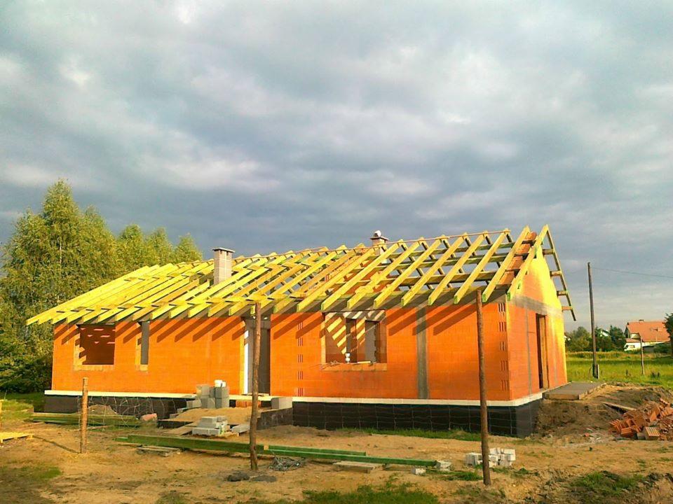 Dom murowany w stanie surowym otwartym z widoczną więźbą dachową, wykonany z pomarańczowych bloczków i izolacją fundamentów, na tle pochmurnego nieba.