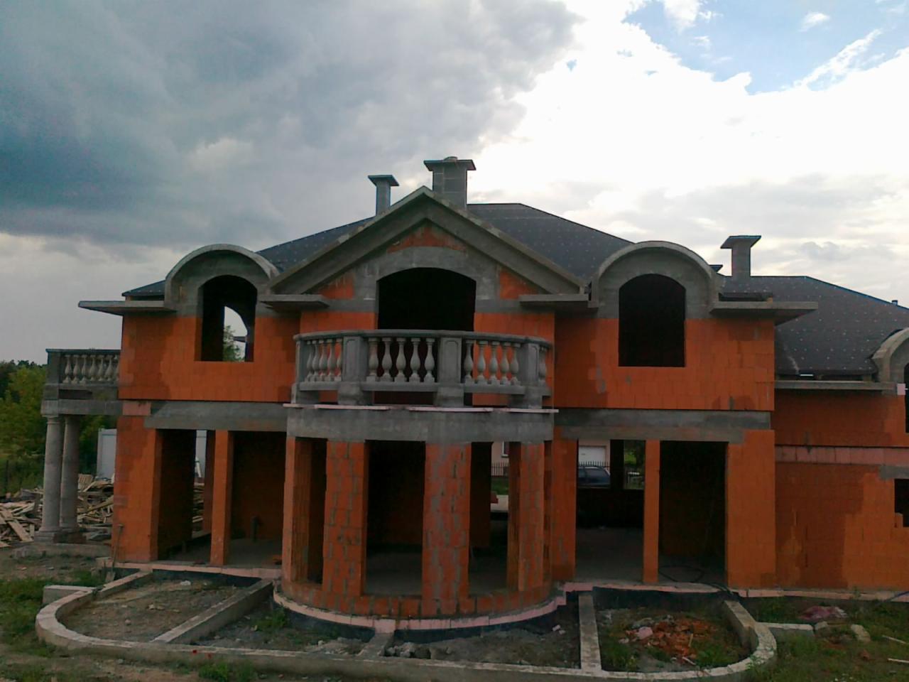 Niedokończony dom z czerwonej cegły z balkonem i widoczną więźbą dachową, otoczony zielenią, pod pochmurnym niebem.