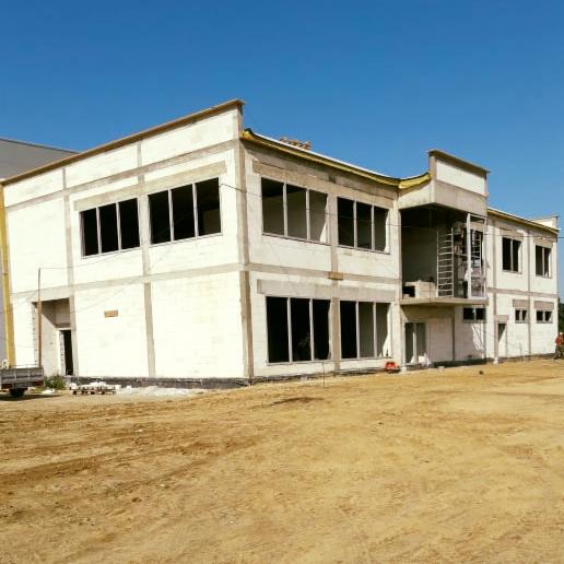 Dwukondygnacyjny budynek w surowym stanie z widocznymi otworami okiennymi i drzwiami, otoczony ziemią, z fragmentem ciężarówki w tle po lewej stronie.