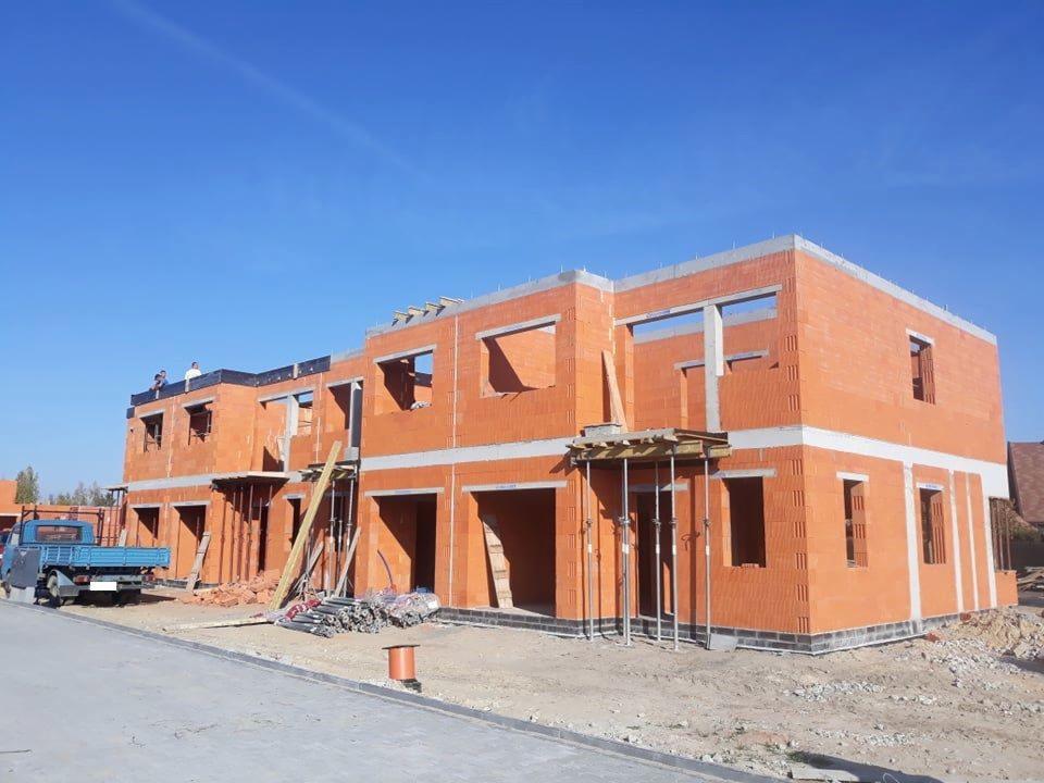 Wznoszenie ścian z czerwonej cegły porotherm w dwukondygnacyjnym budynku w stanie surowym otwartym, widoczni pracownicy na dachu, niebieska ciężarówka w tle.