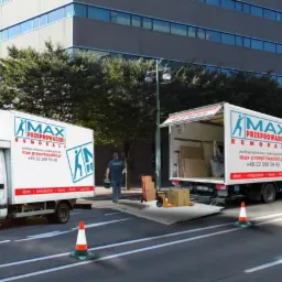 Dwa busy z logo firmy przeprowadzkowej na tle biurowca, rampa rozładunkowa, pracownik w niebieskiej koszulce i kartony.