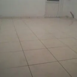 Ułożone jasne, kwadratowe płytki podłogowe z widocznymi fugami w pustym pomieszczeniu. Białe ściany i drzwi w tle.