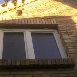 Białe, dwudzielne okno z ceglaną ramą w murze z żółtej cegły, widziane z dołu pod kątem. Częściowo widoczny dach z dachówki.