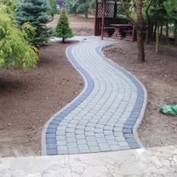 Kręta ścieżka z kostki brukowej w odcieniach szarości i grafitu, obramowana szarym obrzeżem, wijąca się przez ogród z drzewami iglastymi w kierunku drewnianej altany.