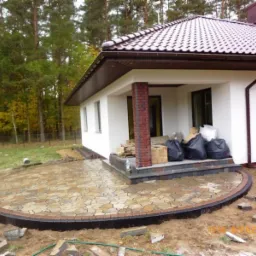 Okrągły taras z nieregularnej kostki w odcieniach beżu i brązu, obrzeżony ciemną kostką, przylegający do białego domu z brązowym dachem na tle drzew.