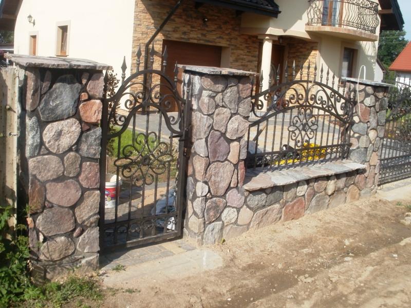 Kamienne słupki i ozdobna, metalowa brama w ogrodzeniu posesji. Widoczna nierówna faktura kamieni i detale zdobień bramy. Uwagę przykuwa solidna konstrukcja i estetyczne wykonanie.
