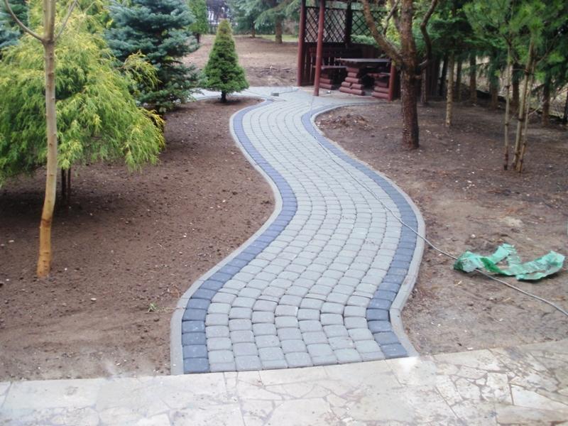 Kręta ścieżka z kostki brukowej w odcieniach szarości i grafitu, obramowana szarym obrzeżem, wijąca się przez ogród z drzewami iglastymi w kierunku drewnianej altany.