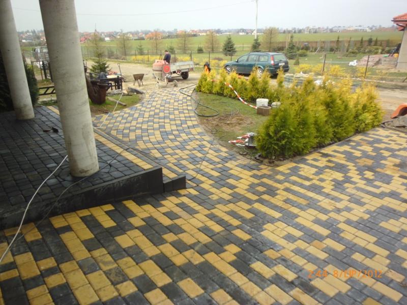 Żółto-szara kostka brukowa ułożona w łukowaty wzór, obok kolumn domu i żywopłotu z tui. W tle betoniarka i krajobraz podmiejski.