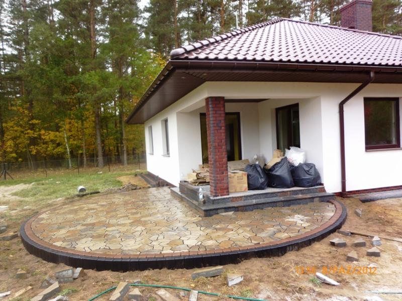 Okrągły taras z nieregularnej kostki w odcieniach beżu i brązu, obrzeżony ciemną kostką, przylegający do białego domu z brązowym dachem na tle drzew.