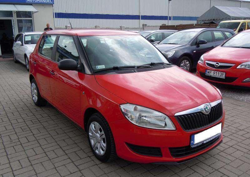 Czerwona Skoda Fabia stoi na placu z kostki brukowej, widok z przodu, inne auta w tle. Na szybie naklejka.