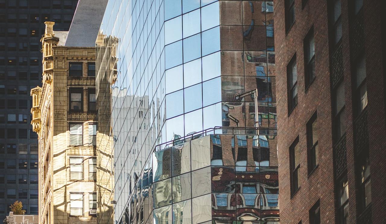 Fasada szklanego biurowca odbija sąsiednie budynki z cegły i kamienia. Geometryczne wzory okien i refleksów tworzą abstrakcyjny kolaż miejski.