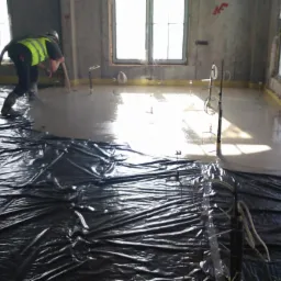 Wylewanie posadzki: robotnik w kamizelce wylewa jasną masę na podłogę, zabezpieczoną czarną folią. Widoczne okna i pionowe znaczniki poziomu.