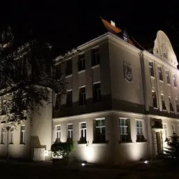 Oświetlona nocą elewacja trzypiętrowego budynku w stylu klasycystycznym z herbem na fasadzie, oświetlenie architektoniczne podkreśla detale budynku.