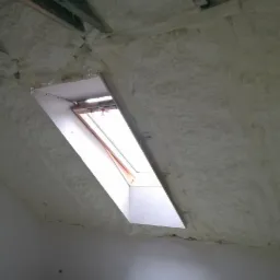 Poddasze po nałożeniu izolacji, widoczne krokwie i okno dachowe, pianka pokrywa całą powierzchnię, nierównomierna struktura, na górze widoczne elementy konstrukcji dachu.
