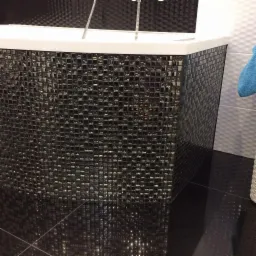 Błyszcząca, czarna mozaika z małych kwadratów zdobi obudowę wanny w nowoczesnej łazience, z ciemną, lustrzaną podłogą i fragmentem białej, strukturalnej ściany w tle.