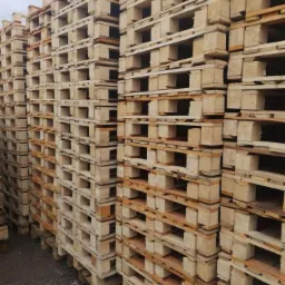Stosy jasnych, surowych palet drewnianych ułożonych jeden na drugim, widoczne ślady użytkowania na niektórych elementach, szare tło podłoża.