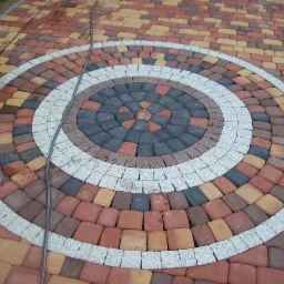 Kolorowa kostka brukowa ułożona w okrągły wzór z szarym obramowaniem, widok z góry, kabel na powierzchni. Różnokolorowe odcienie czerwieni, brązu i szarości.