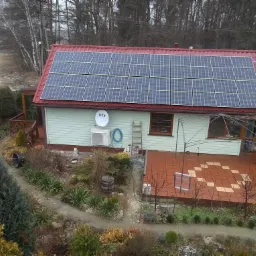 Dom jednorodzinny z czerwonym dachem, na którym zamontowano panele słoneczne. Na ścianie budynku widoczna antena satelitarna i klimatyzator. W tle drzewa i pochmurne niebo.