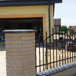 Nowoczesne ogrodzenie z elementami z kostki brukowej, czarne metalowe przęsła, na tle domu z ciemną dachówką i żółtą elewacją. Słoneczny dzień.