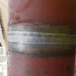 Czerwony metalowy słup z widocznym, świeżym spawem o charakterystycznym wzorze łusek, wykonanym techniką TIG, na tle rozmytego otoczenia budowlanego.